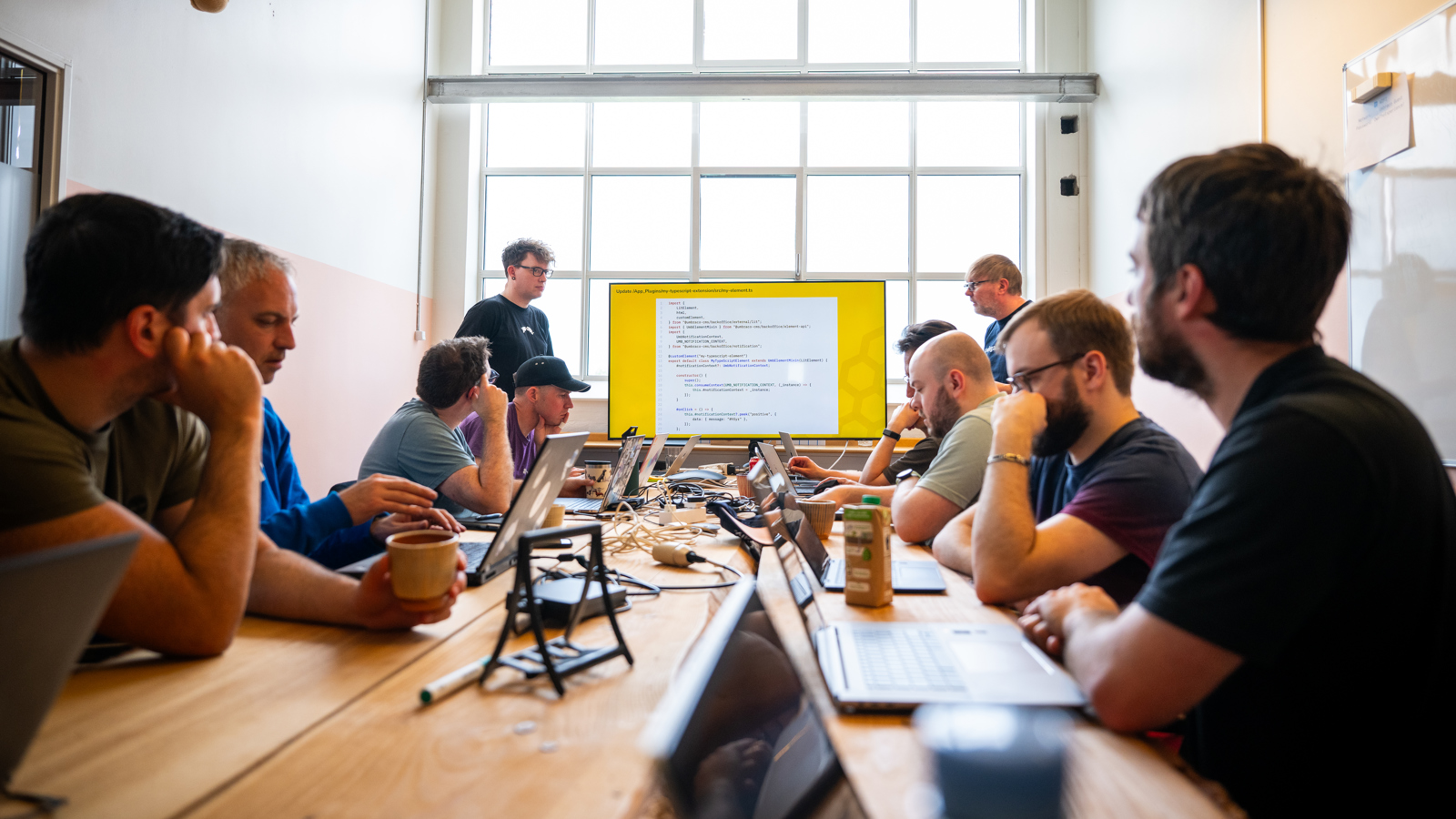 A group of people are sitting around a long wooden table in a well-lit room with a large window in the background. They are engaged in a meeting or workshop, with laptops and notebooks in front of them. Two individuals are standing at the front of the room, presenting a slide on a large screen that displays code. The attendees appear focused and attentive, suggesting a technical or coding workshop. The room has a modern industrial look with exposed ducts on the ceiling. Click to open full size image.