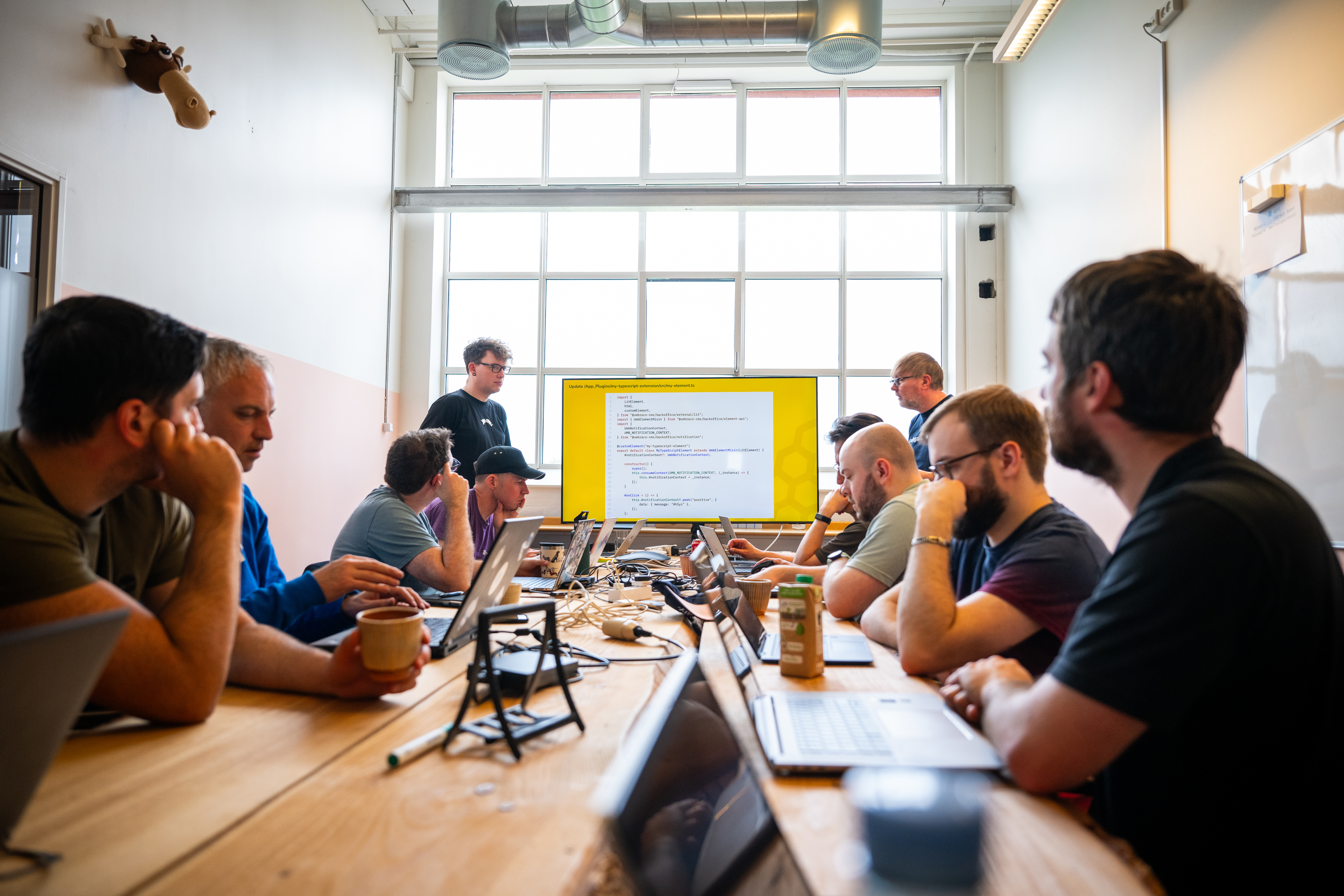 A group of people are sitting around a long wooden table in a well-lit room with a large window in the background. They are engaged in a meeting or workshop, with laptops and notebooks in front of them. Two individuals are standing at the front of the room, presenting a slide on a large screen that displays code. The attendees appear focused and attentive, suggesting a technical or coding workshop. The room has a modern industrial look with exposed ducts on the ceiling.