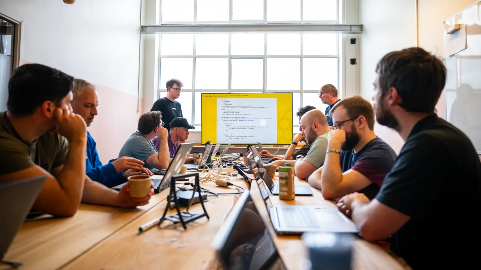 Click to open full size image. A group of people are sitting around a long wooden table in a well-lit room with a large window in the background. They are engaged in a meeting or workshop, with laptops and notebooks in front of them. Two individuals are standing at the front of the room, presenting a slide on a large screen that displays code. The attendees appear focused and attentive, suggesting a technical or coding workshop. The room has a modern industrial look with exposed ducts on the ceiling. Click to open full size image.