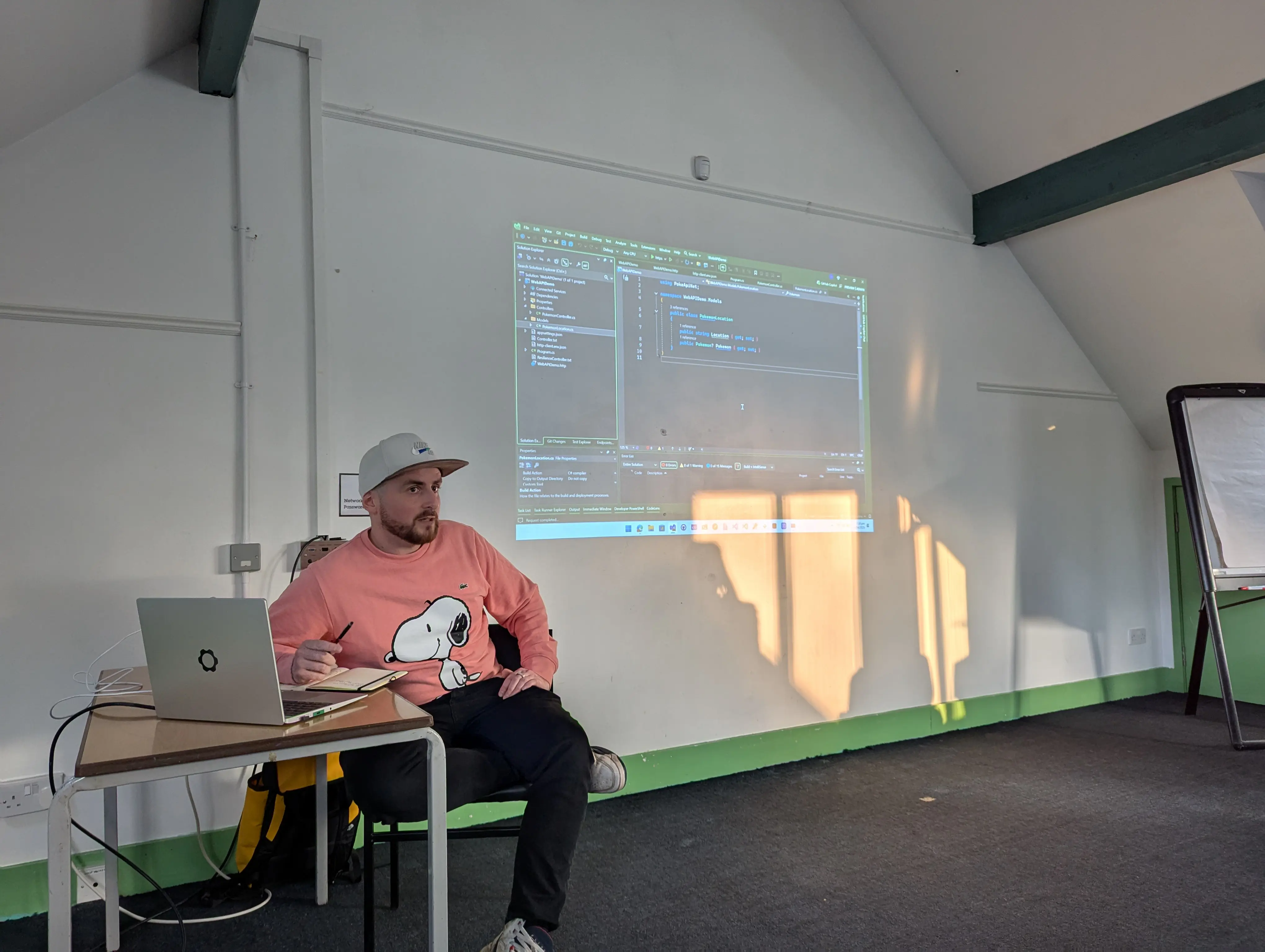 A speaker (Luke) sits in front of a projected slide at the umBristol April Umbraco meetup, smiling while presenting. He wears a pink snoopy hoodie and dark trousers. A laptop sits on a small table nearby, connected to the projector. The room has a sloped ceiling and green trim along the walls.