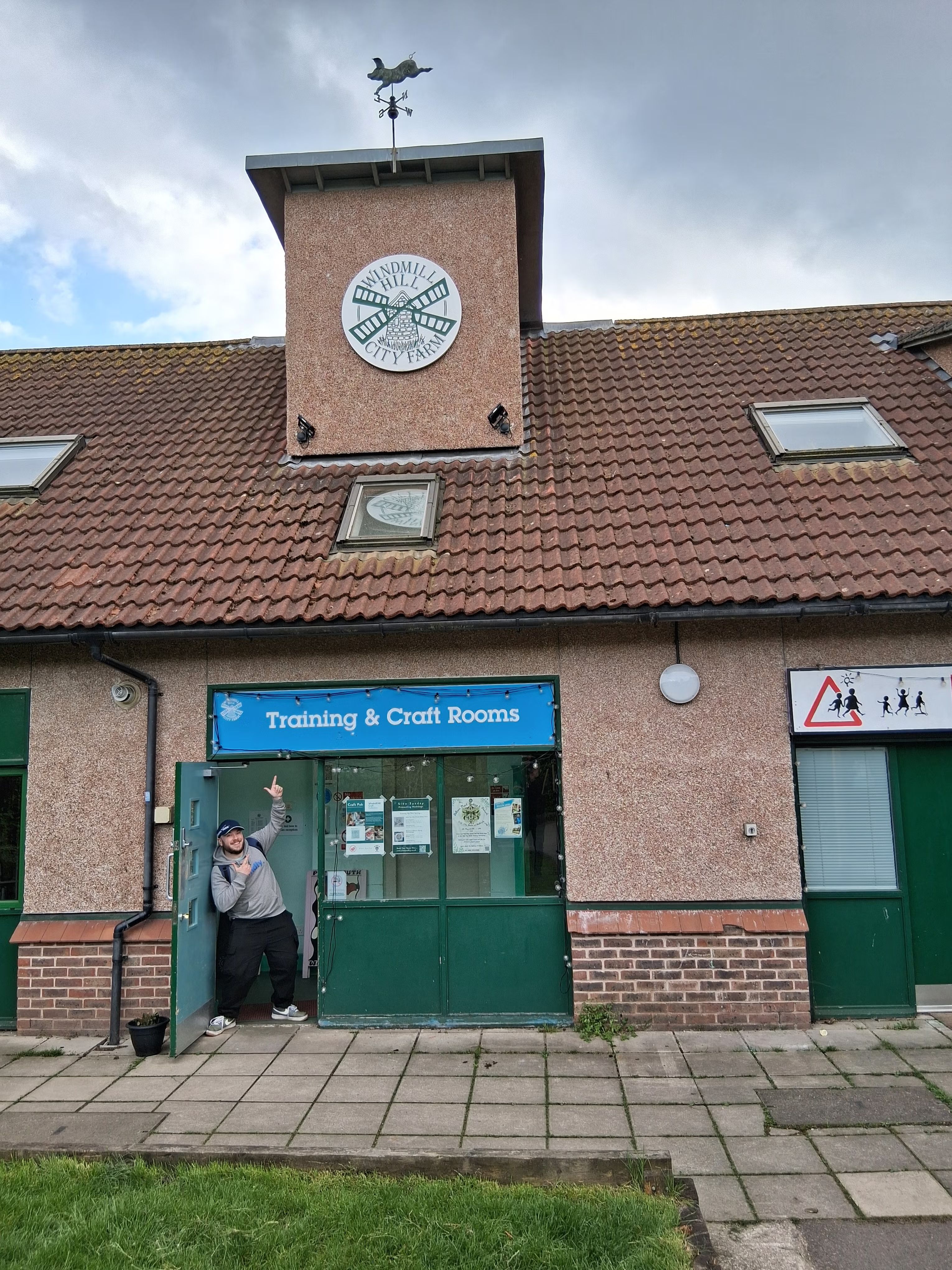 Owain standing in the doorway of a building, pointing and smiling at a sign that says "Training & Craft rooms" Click to open full size image.