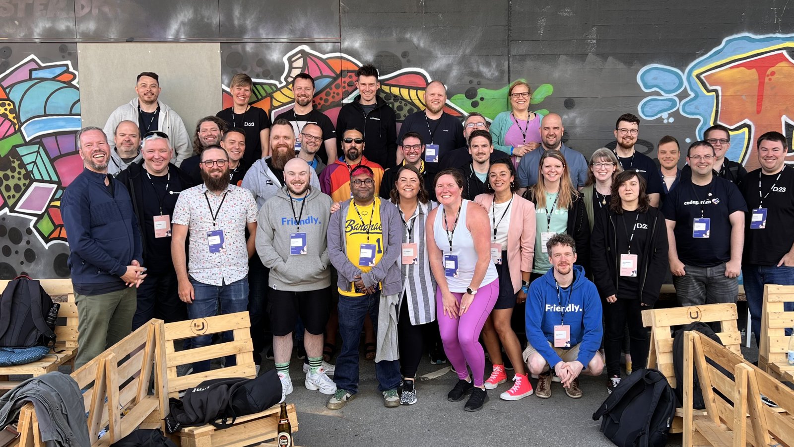 A group photo of CODECABIN attendees taken at the Codegarden conference. The group in the phot consists of around 33 people, standing and seated in an outdoor setting against a wall adorned with colorful graffiti art. The attendees are smiling and appear to be in high spirits, many wearing conference lanyards and badges. They are casually dressed, with some wearing branded t-shirts and hoodies. The seating area is made up of wooden pallet benches. The atmosphere is vibrant and energetic. Click to open full size image.