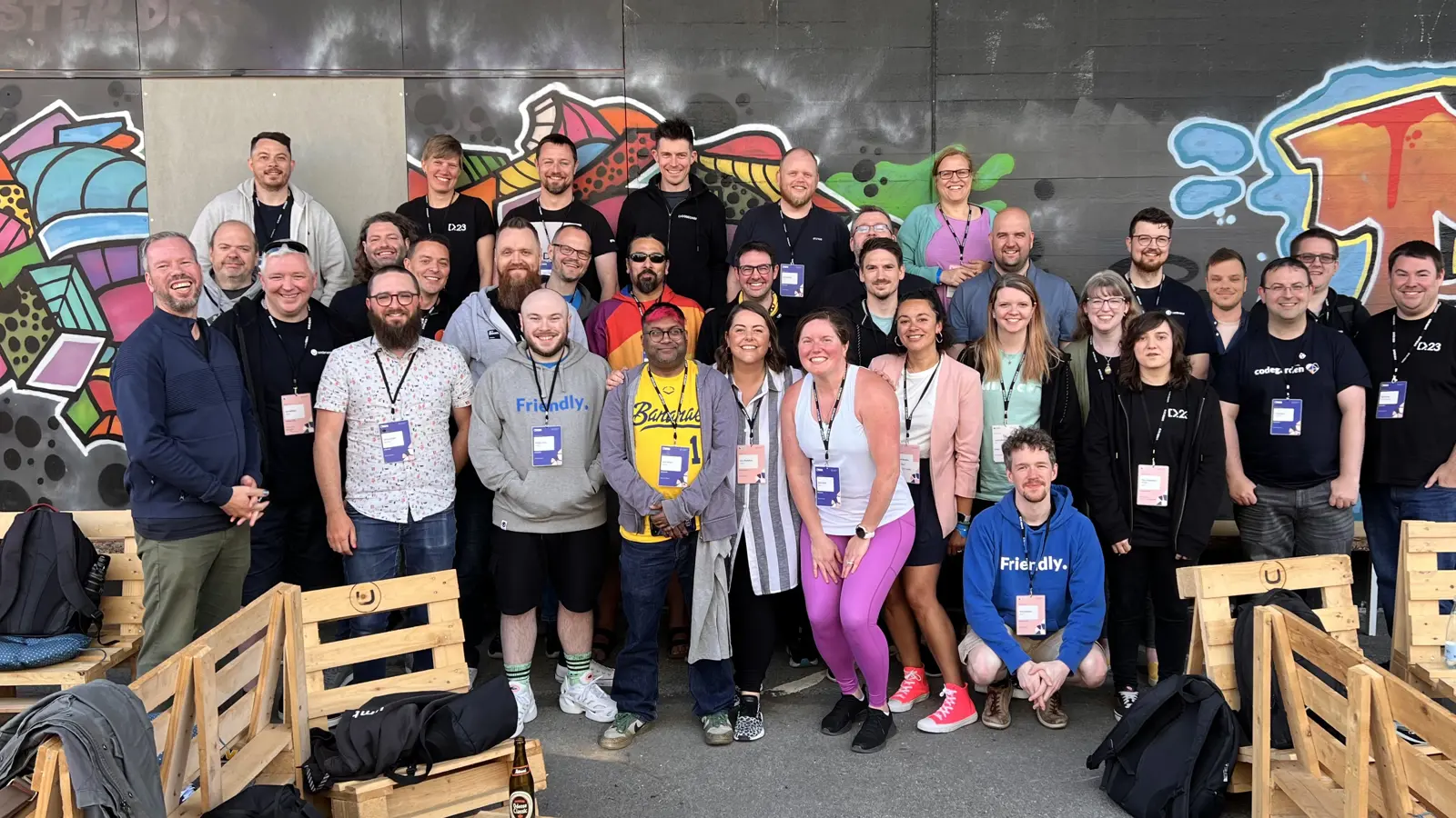 Click to open full size image. A group photo of CODECABIN attendees taken at the Codegarden conference. The group in the phot consists of around 33 people, standing and seated in an outdoor setting against a wall adorned with colorful graffiti art. The attendees are smiling and appear to be in high spirits, many wearing conference lanyards and badges. They are casually dressed, with some wearing branded t-shirts and hoodies. The seating area is made up of wooden pallet benches. The atmosphere is vibrant and energetic. Click to open full size image.