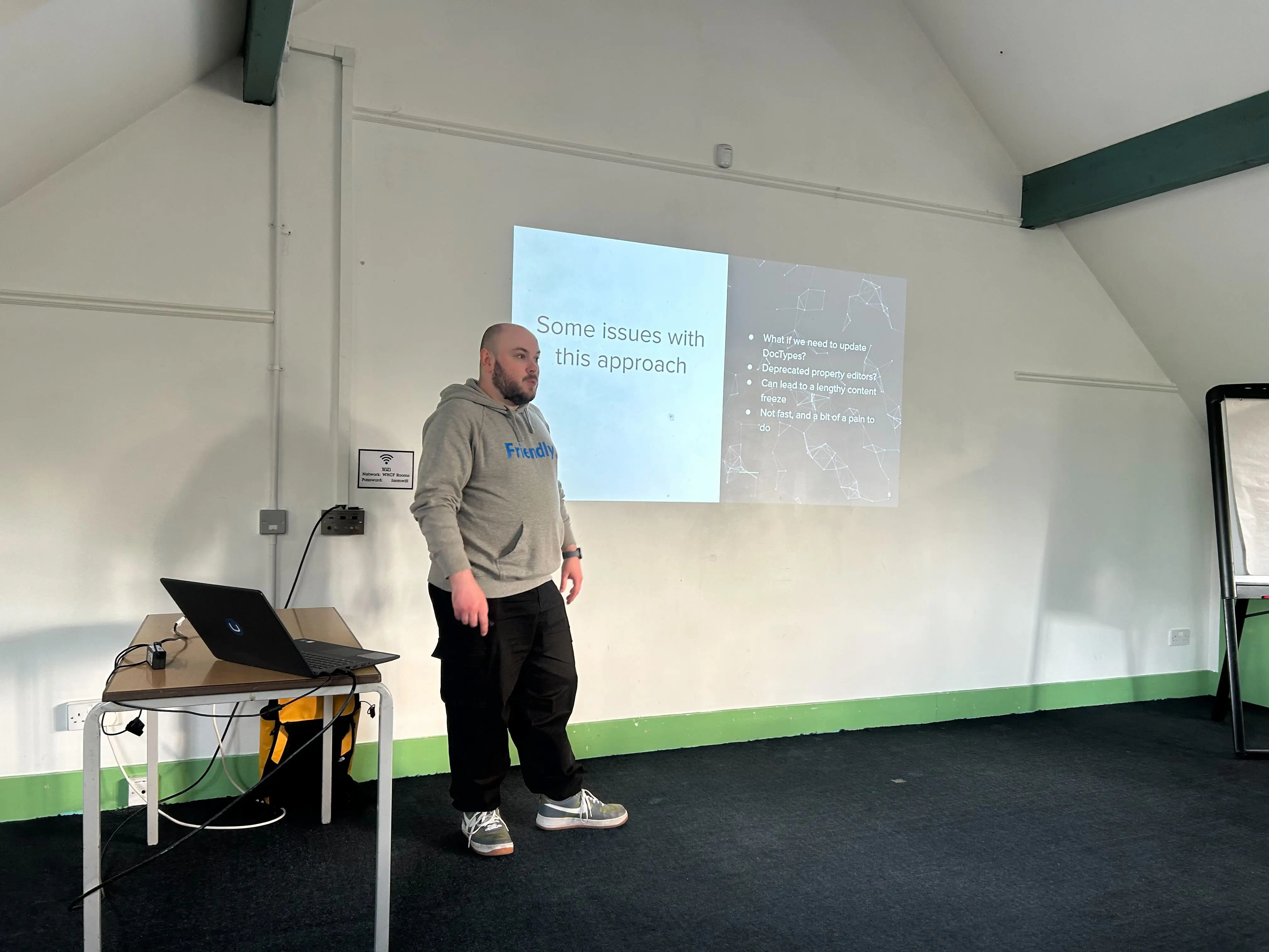 A speaker (Owain) stands in front of a projected slide at the umBristol April Umbraco meetup, smiling while presenting. He wears a grey 'Friendly' hoodie and dark trousers. A laptop sits on a small table nearby, connected to the projector. The room has a sloped ceiling and green trim along the walls.