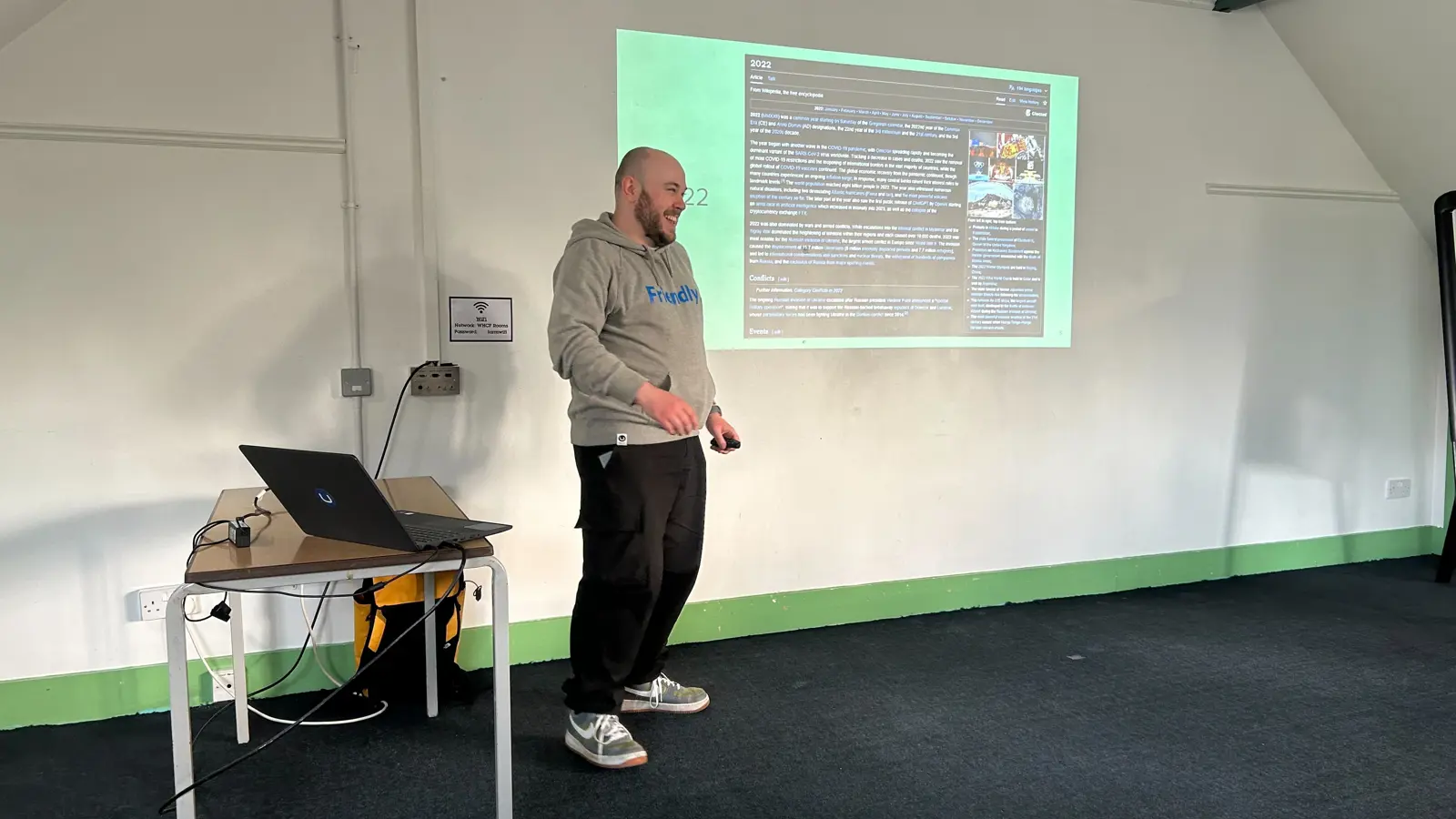 A speaker (Owain) stands in front of a projected slide at the umBristol April Umbraco meetup, smiling while presenting. He wears a grey 'Friendly' hoodie and dark trousers. A laptop sits on a small table nearby, connected to the projector. The room has a sloped ceiling and green trim along the walls. Click to open full size image.