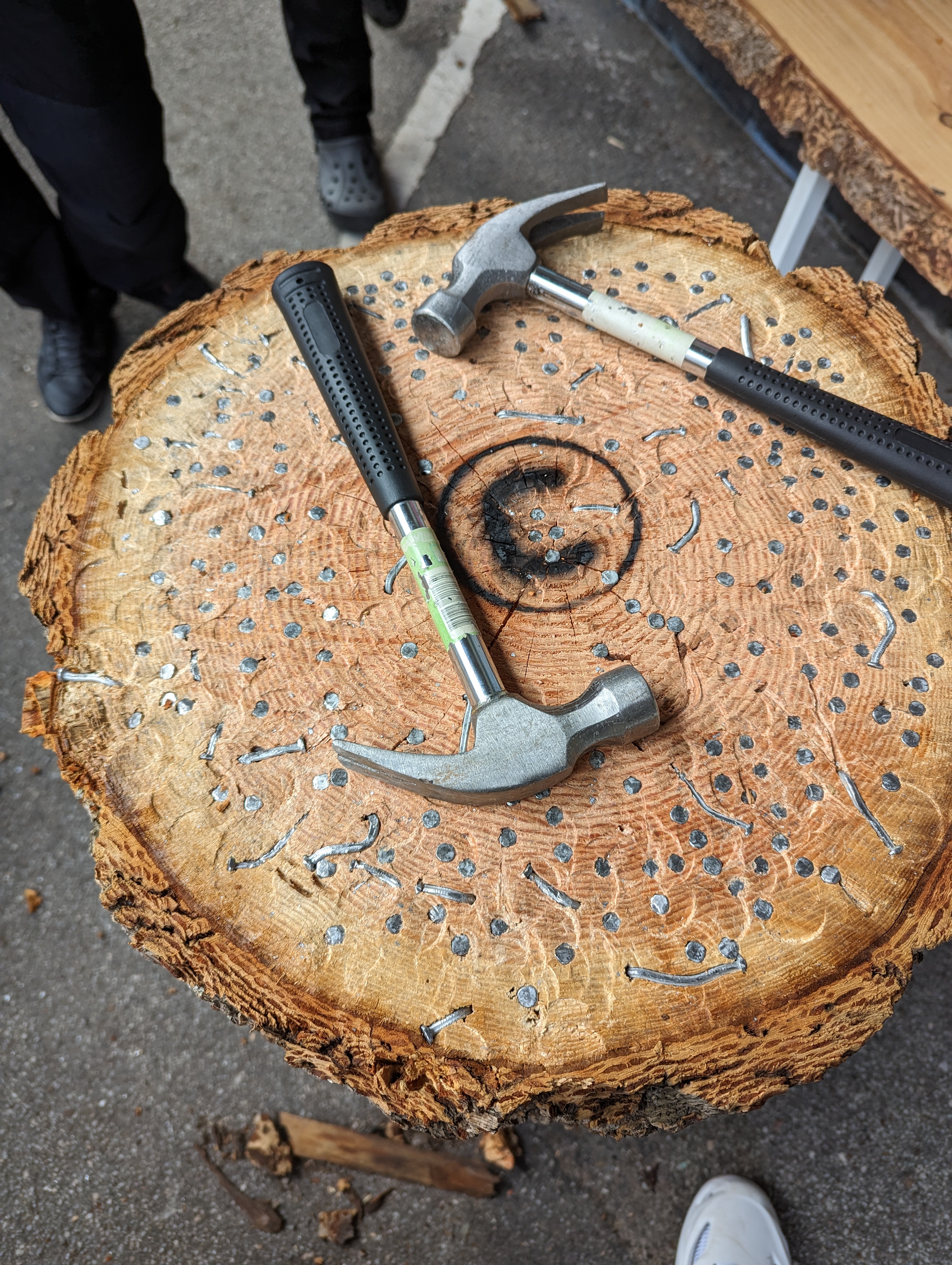  A close-up view of a tree stump used for a hammering game, featuring two hammers lying on top. The surface of the stump is embedded with numerous nails, some hammered in and others bent or partially driven in, with the Umbraco logo burned into the centre. The stump's outer bark is rough and textured, contrasting with the smoother, nail-filled interior. The ground around the stump is paved, and the lower legs and feet of some individuals are visible in the background, suggesting an outdoor setting.