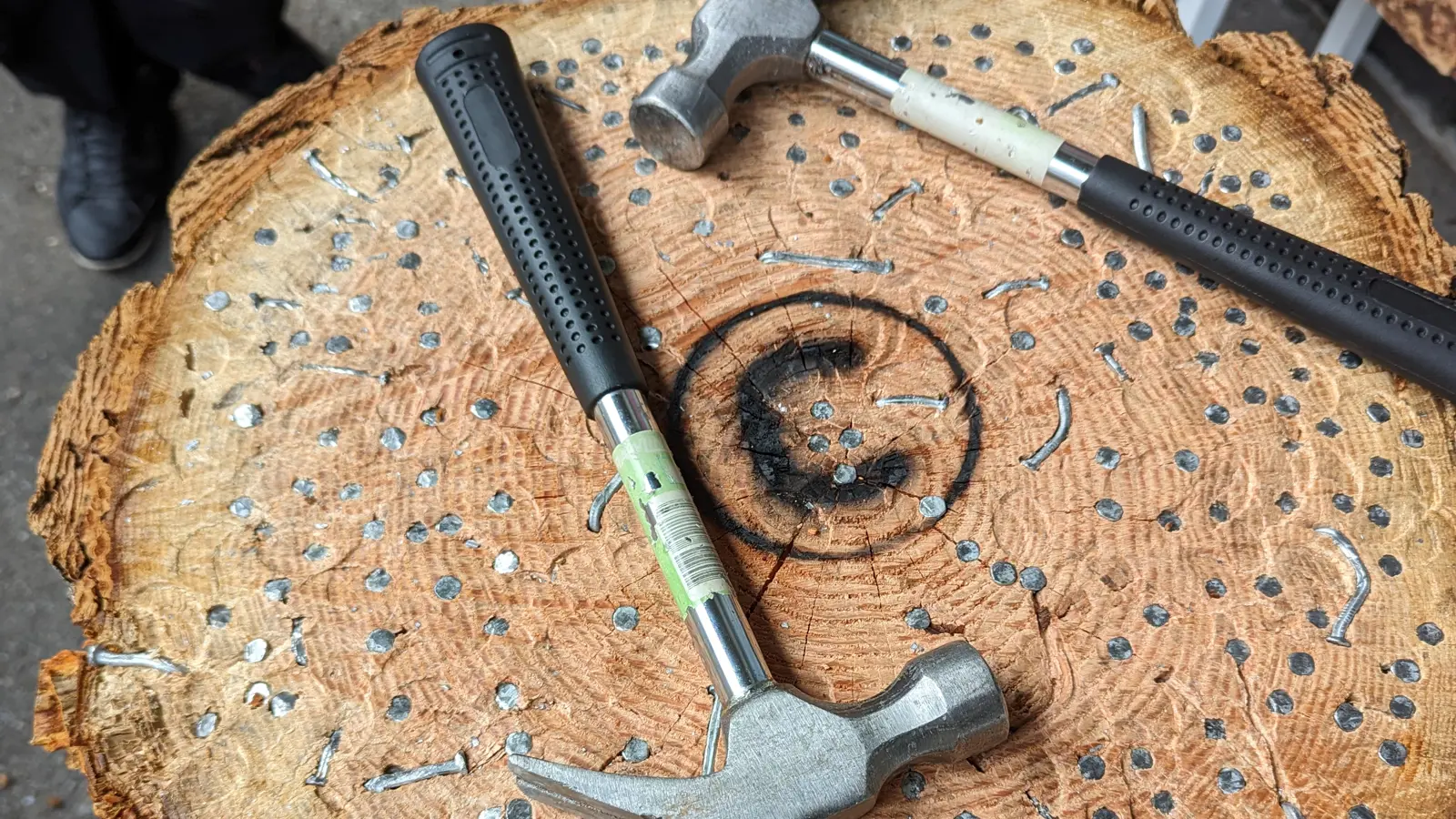 Click to open full size image.  A close-up view of a tree stump used for a hammering game, featuring two hammers lying on top. The surface of the stump is embedded with numerous nails, some hammered in and others bent or partially driven in, with the Umbraco logo burned into the centre. The stump's outer bark is rough and textured, contrasting with the smoother, nail-filled interior. The ground around the stump is paved, and the lower legs and feet of some individuals are visible in the background, suggesting an outdoor setting. Click to open full size image.