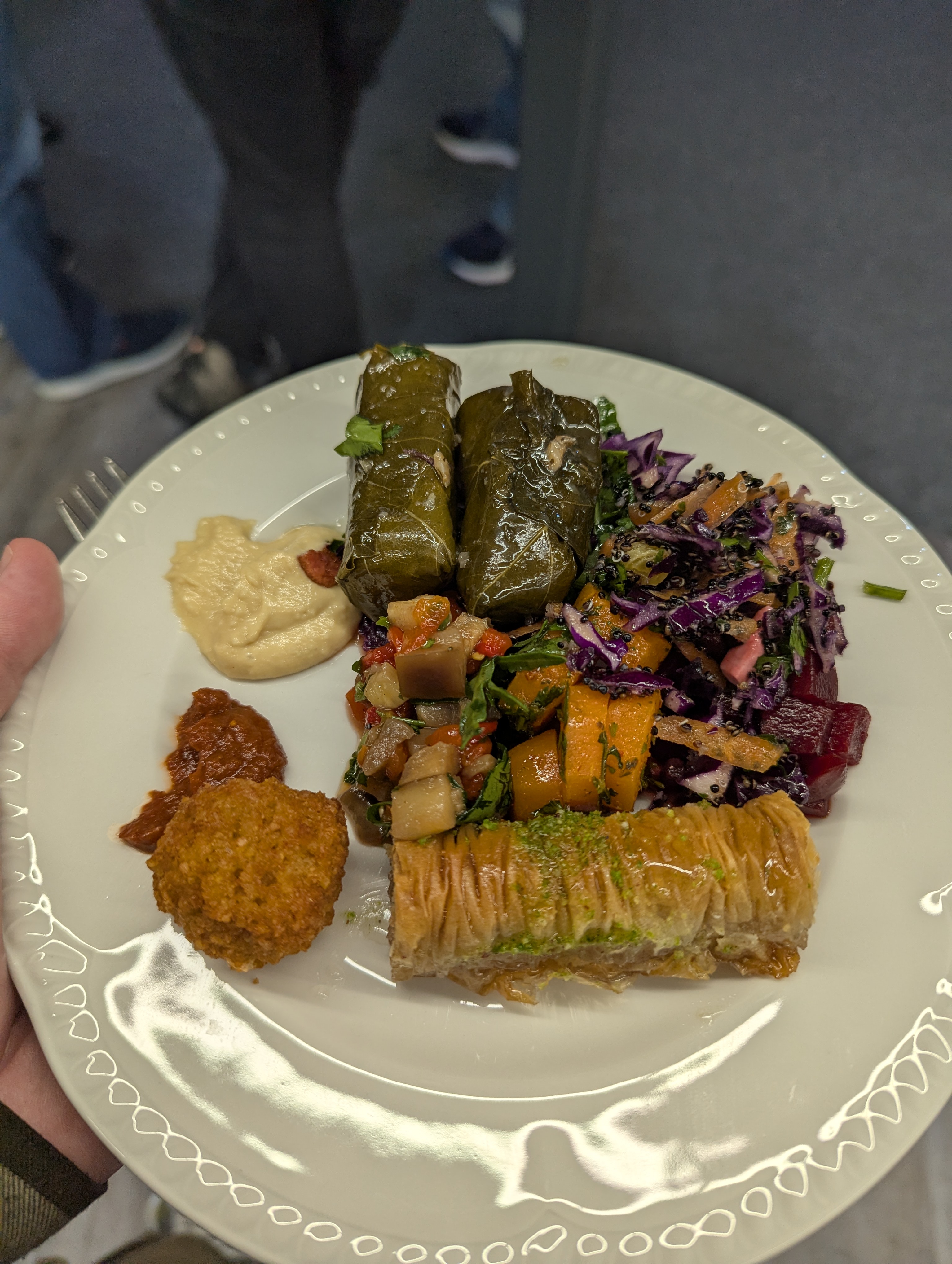 A close-up of a plate of Greek food at the DDD South West conference, featuring stuffed vine leaves, baklava, a colorful salad with vegetables, hummus, a small falafel ball, and a spicy red sauce. Click to open full size image.
